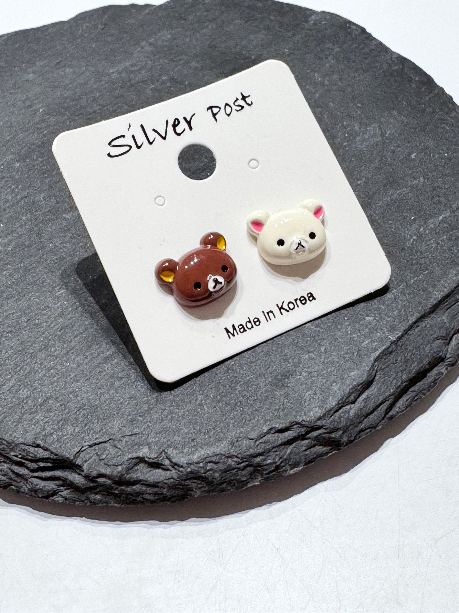 A pair of mismatched brown and ivory bear stud earrings on a Silver Post card, placed on a black slate tray.