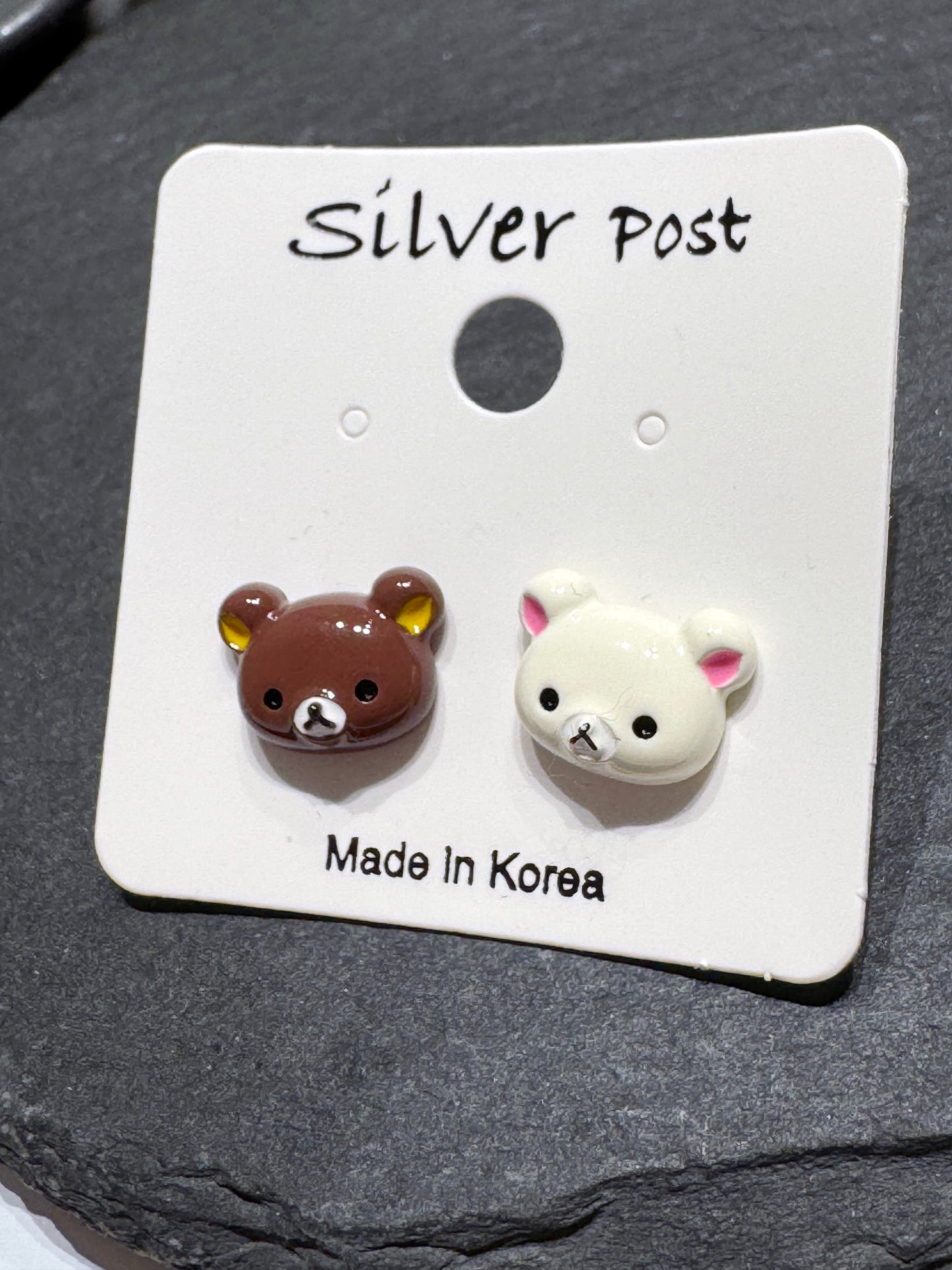 A pair of mismatched brown and ivory bear stud earrings on a Silver Post card, placed on a black slate tray.
