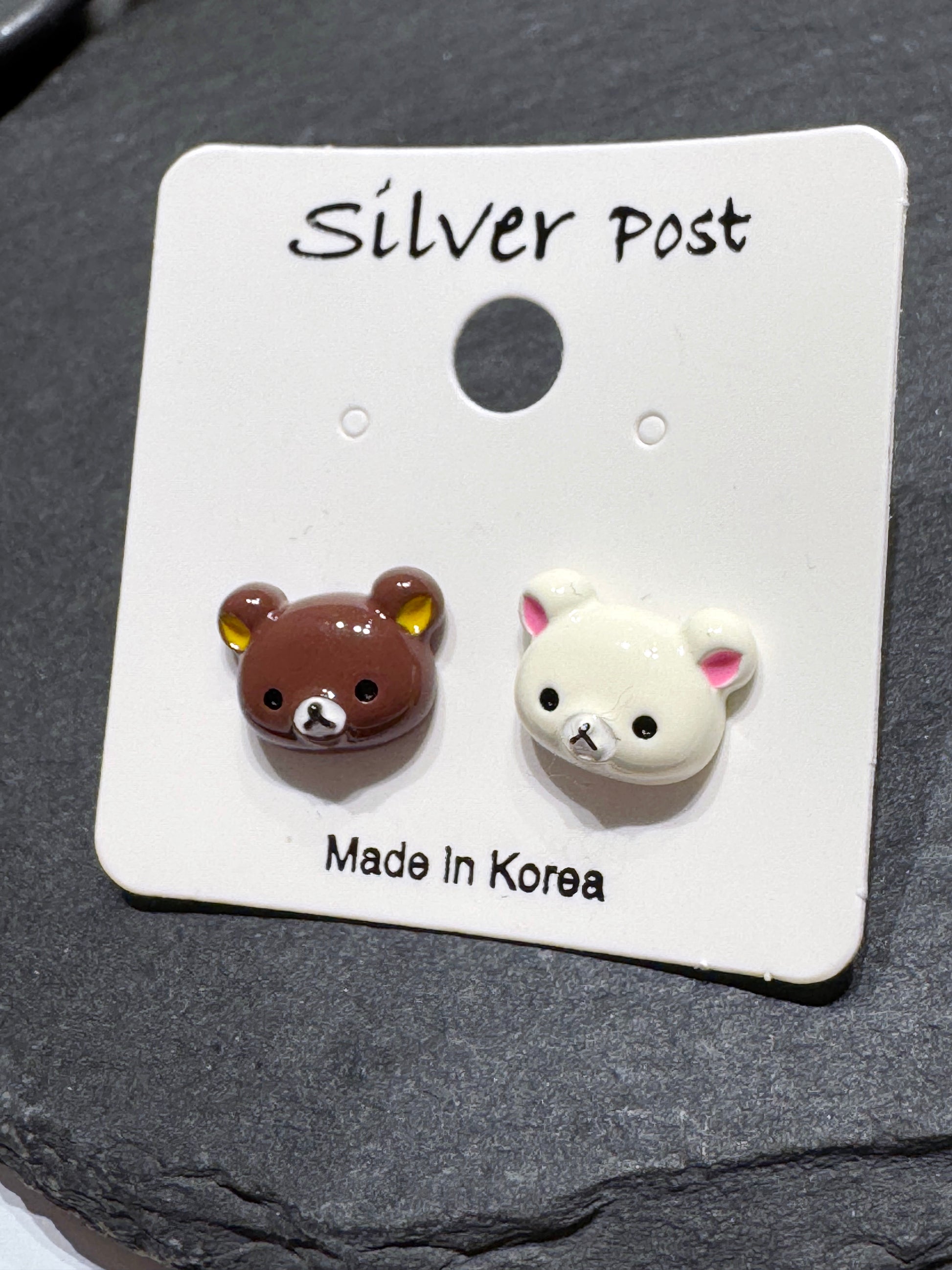 A pair of mismatched brown and ivory bear stud earrings on a Silver Post card, placed on a black slate tray.