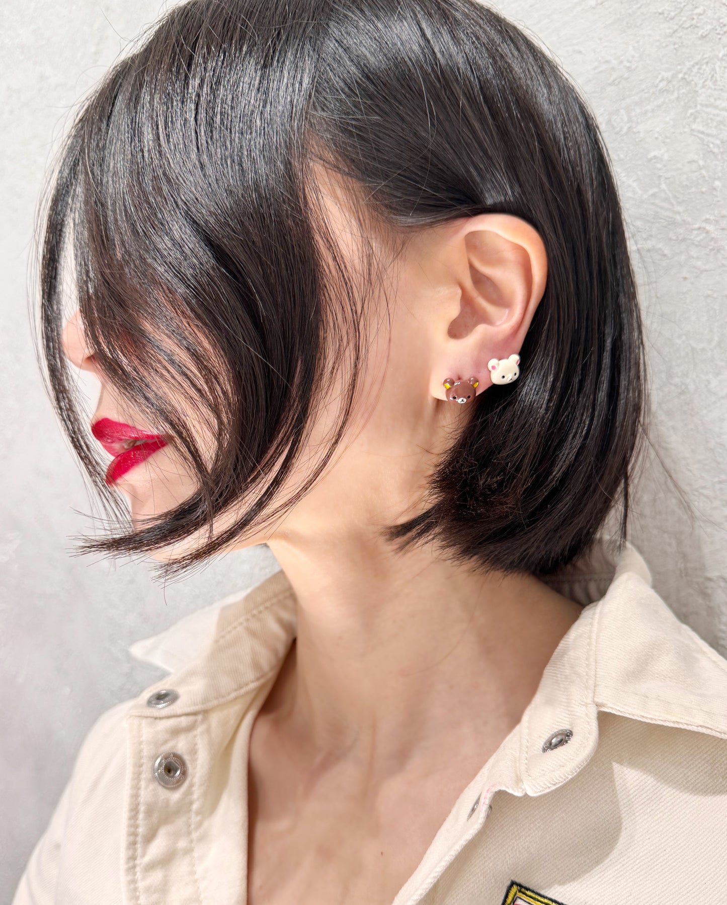 Artistic close-up of mismatched bear-head stud earrings worn on the earlobe, with short black hair partially covering the face.