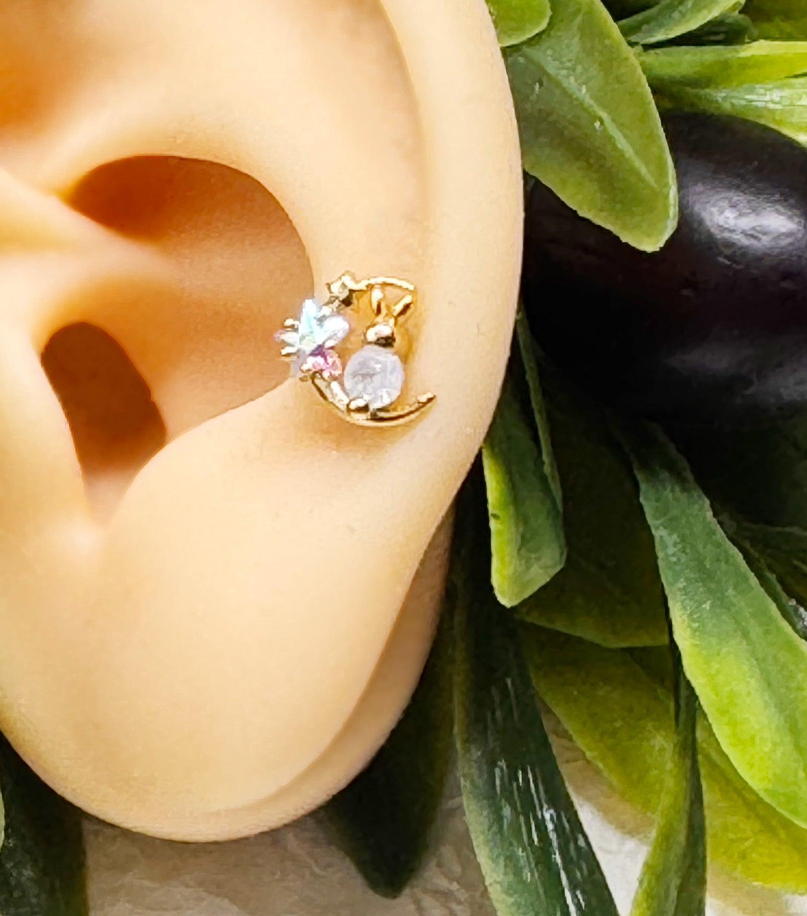 Close-up of a gold-plated bunny and moon barbell earring styled on a faux ear with green leaves in the background. The earring features cubic stars and a round central gem.