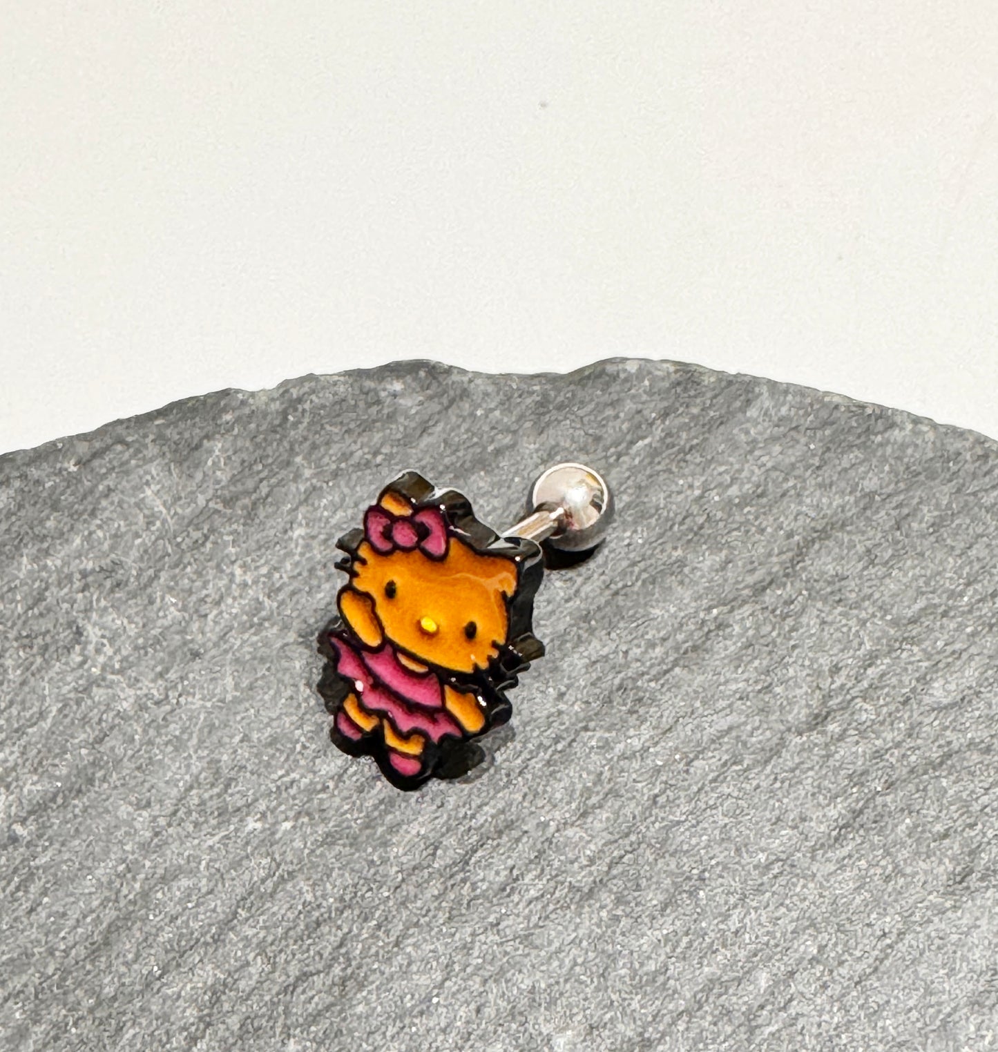 Adorable Hello Kitty–inspired piercing featuring a playful Hawaii-style cat in pink or red dress. Made with hypoallergenic surgical steel. Sold as single.