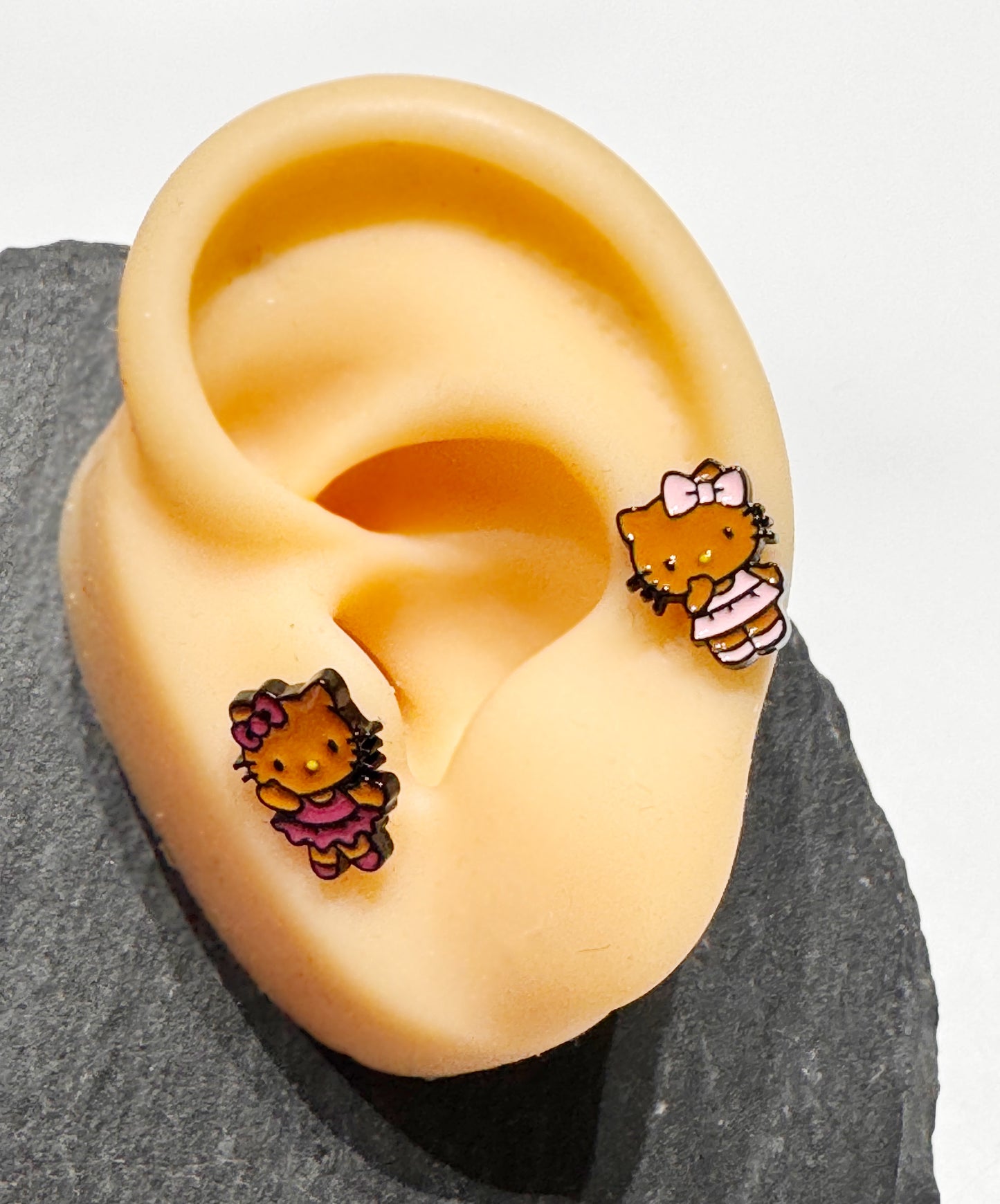 Adorable Hello Kitty–inspired piercing featuring a playful Hawaii-style cat in pink or red dress. Made with hypoallergenic surgical steel. Sold as single.