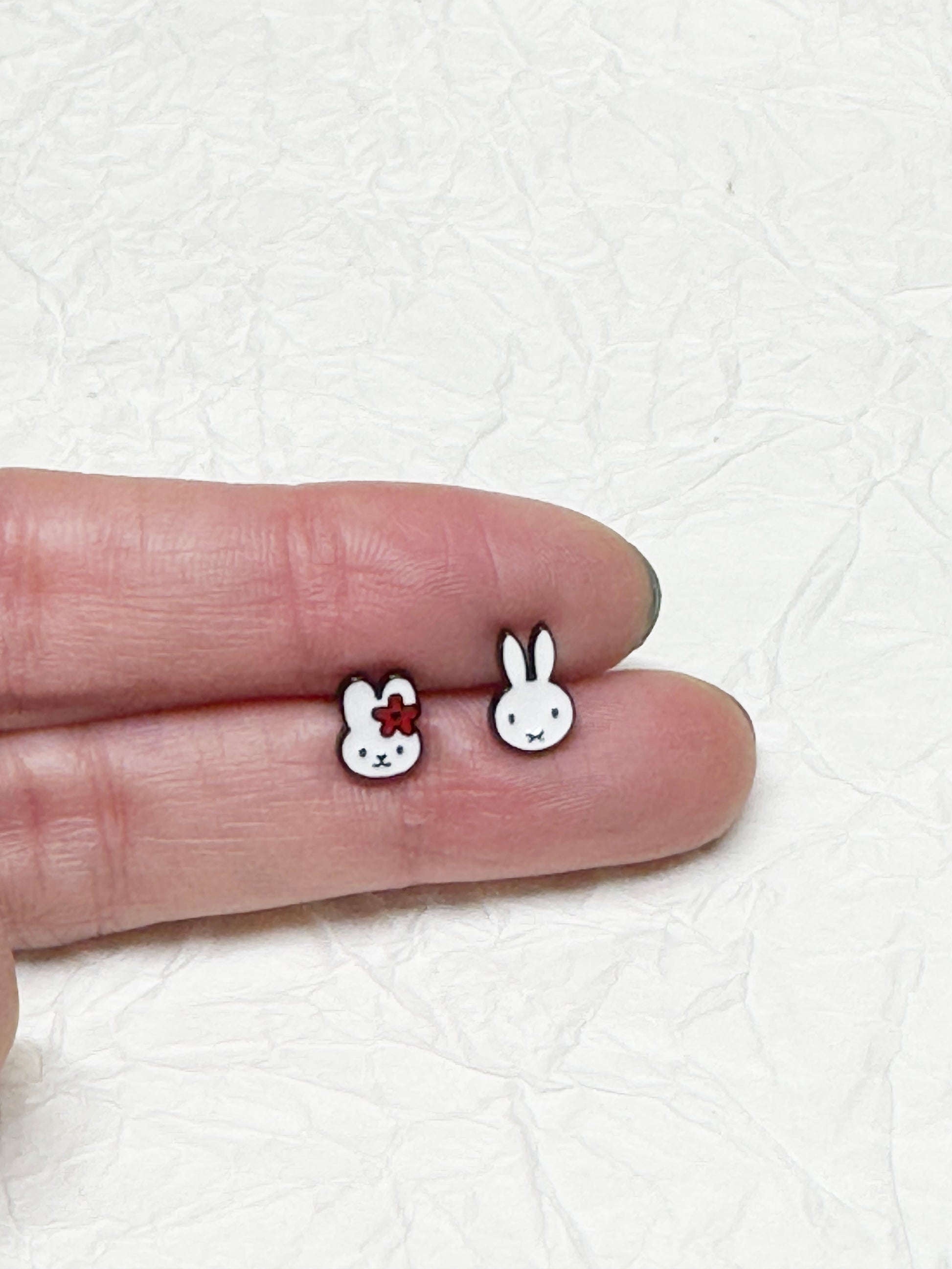 Miffy character earrings shown on fingertips for size comparison; one has a red flower, the other is a classic outline — placed over a textured white paper background.