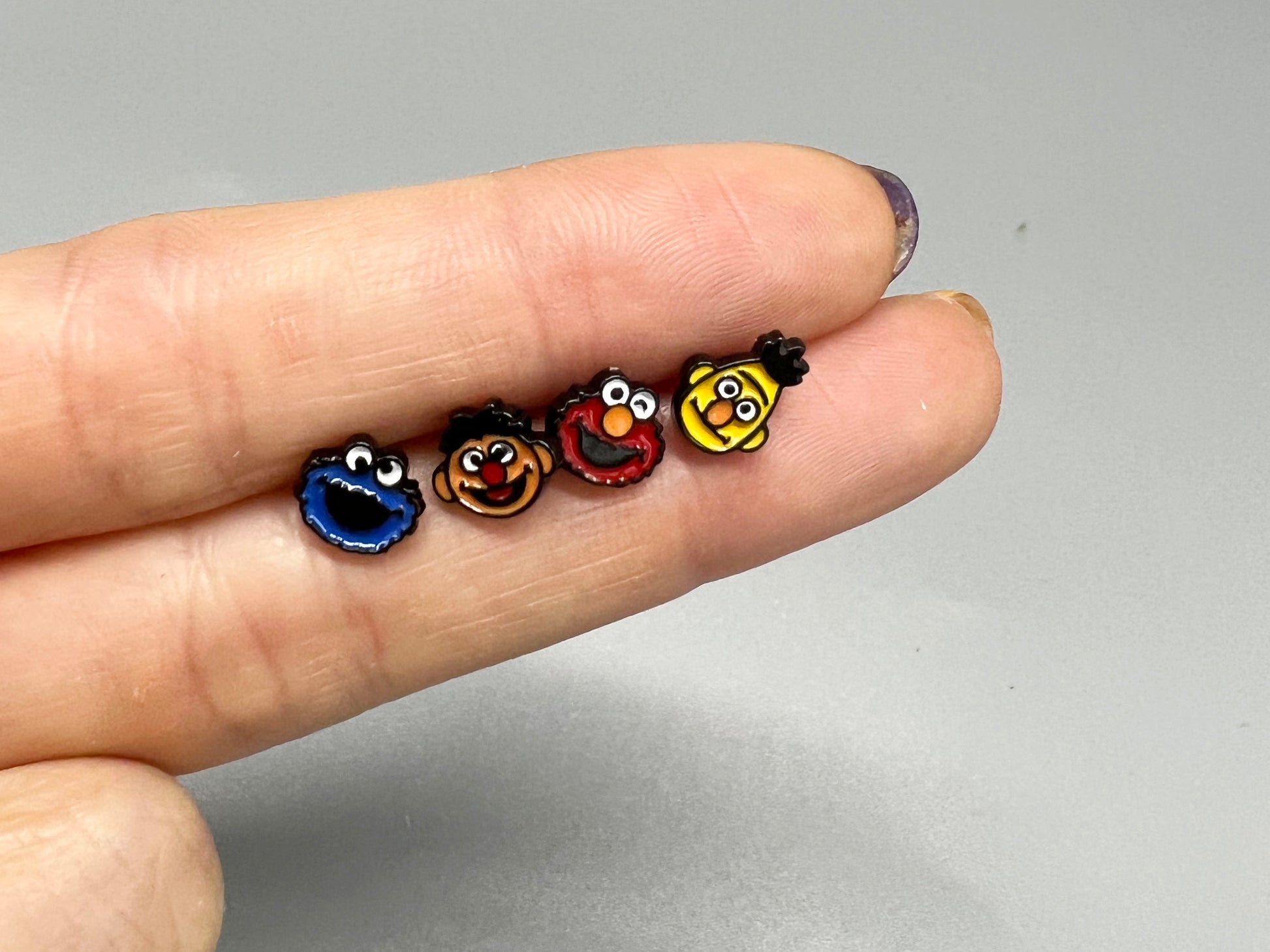 Hand holding four mini Sesame Street character earrings showing compact size and colorful enamel details