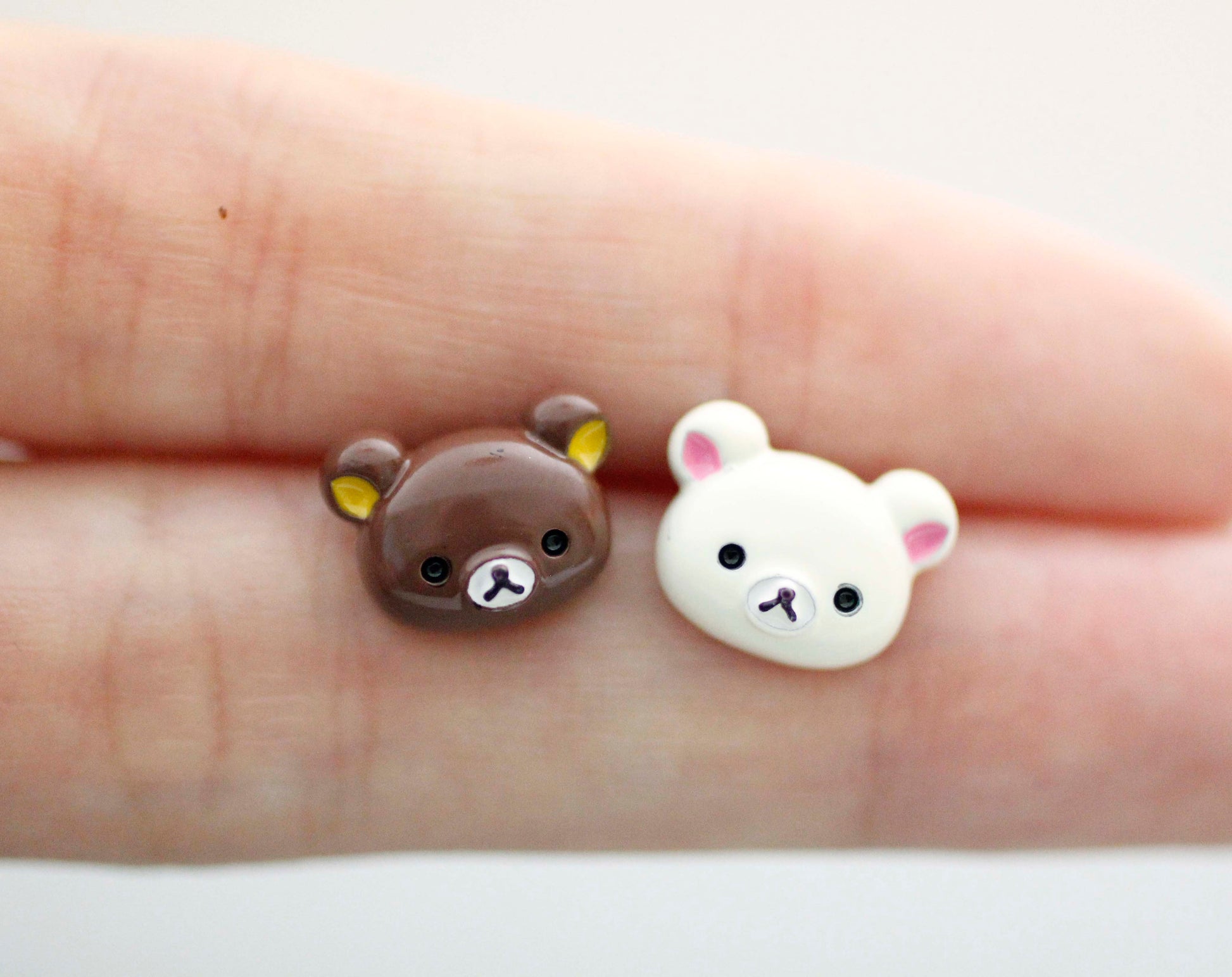 Close-up of two tiny handmade bear-head stud earrings, one brown and one ivory, resting on a person’s fingertip. Silver post, made in Korea.