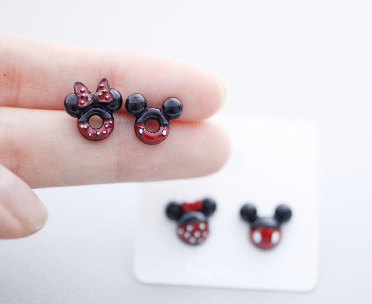 Size comparison Mickey donut and Minnie ladybug earrings on finger approximately 1cm