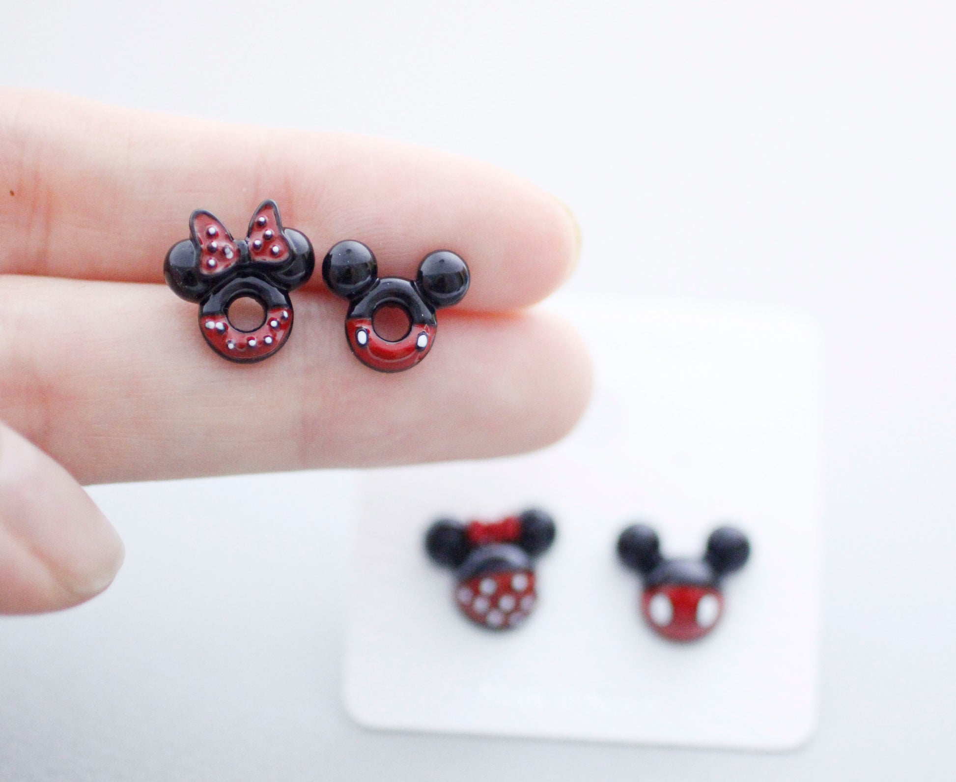 Size comparison Mickey donut and Minnie ladybug earrings on finger approximately 1cm
