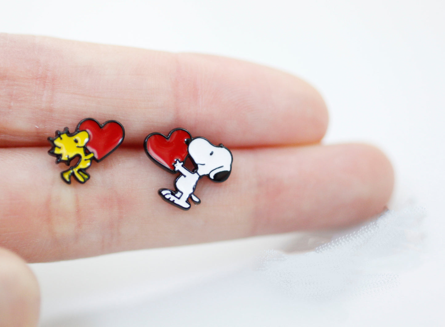Close-up of Snoopy and Woodstock holding red hearts, enamel barbell earrings shown on fingertips.