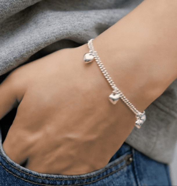 Close-up of a dainty 925 silver heart charm bracelet worn on a wrist against beige linen top and blue denim jeans, casual everyday layered style