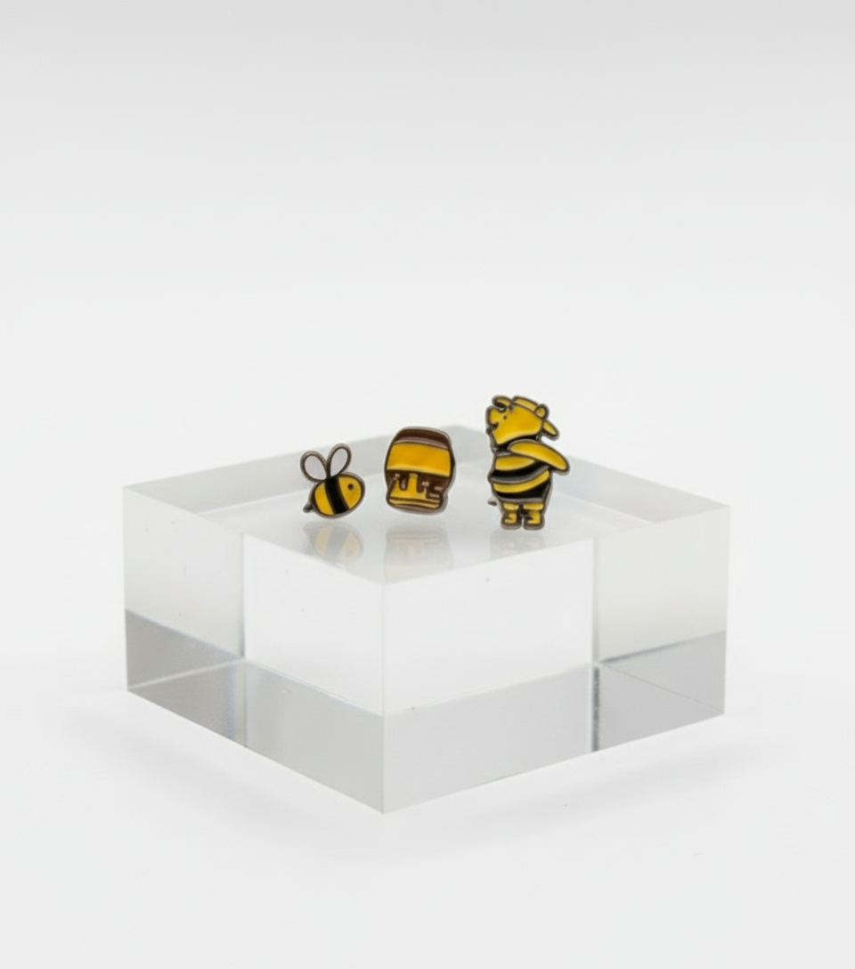 Bee, honey pot, and Winnie the Pooh earrings standing on a clear acrylic block with a blurred Pooh and Tigger figure in the background.
