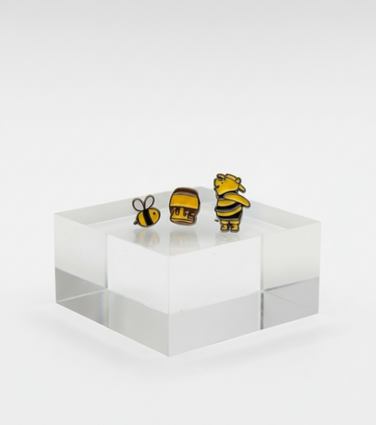 Bee, honey pot, and Winnie the Pooh earrings standing on a clear acrylic block with a blurred Pooh and Tigger figure in the background.