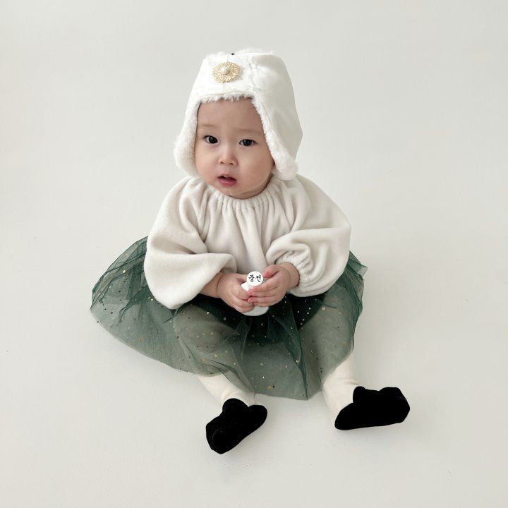 Baby sitting in a cream outfit and forest green tulle skirt, wearing a white fuzzy bonnet with gold brooch.