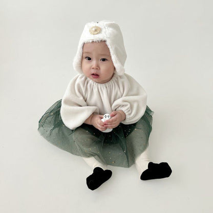 Baby sitting in a cream outfit and forest green tulle skirt, wearing a white fuzzy bonnet with gold brooch.
