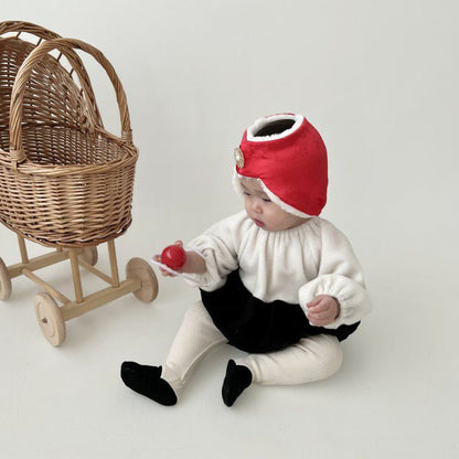 Side view of a baby in a red Korean traditional bonnet (jobawi), holding a red heart toy, sitting next to a wooden basket