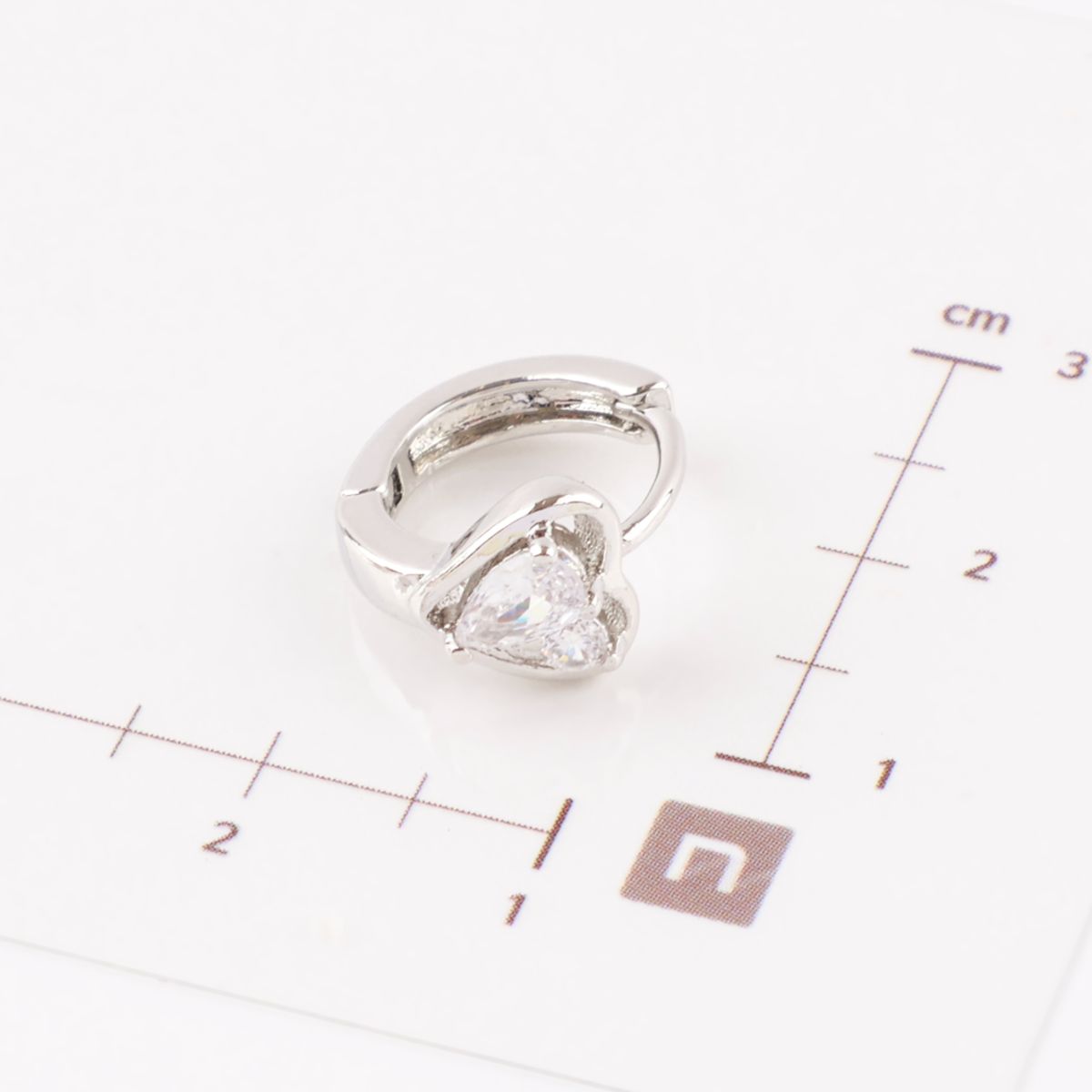 Silver heart huggie hoop earring placed on a white background with centimeter scale showing size reference.