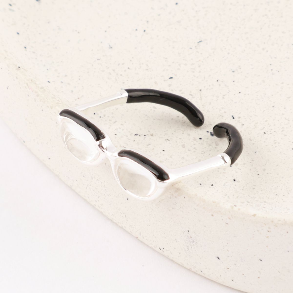 Flat lay of silver spectacle ring showing side view with black enamel temple tips on white stone surface