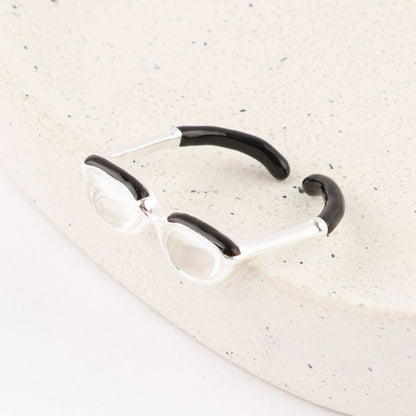 Flat lay of silver spectacle ring showing side view with black enamel temple tips on white stone surface