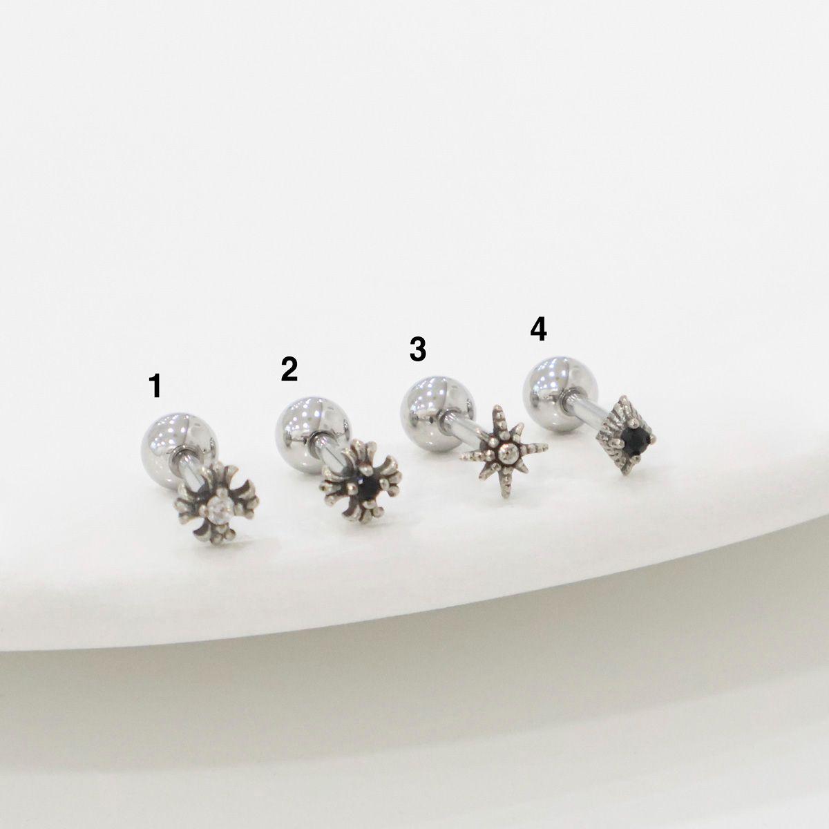 Four stainless steel barbell piercings laid out in a row, numbered from 1 to 4. Each has a unique charm: crystal burst, black sun, radiant star, and gothic square. Product photography on white ceramic plate.