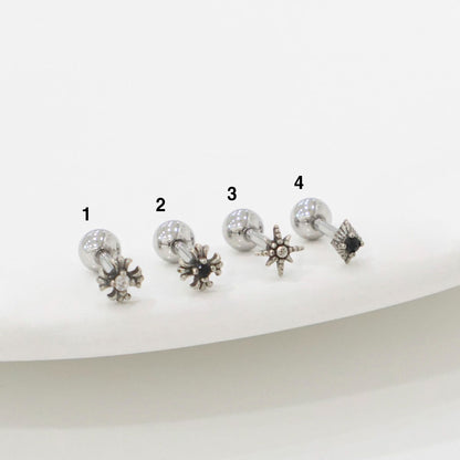 Four stainless steel barbell piercings laid out in a row, numbered from 1 to 4. Each has a unique charm: crystal burst, black sun, radiant star, and gothic square. Product photography on white ceramic plate.
