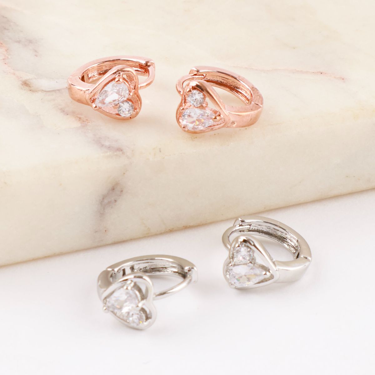 Close-up of rose gold and silver mini huggie hoop earrings with heart-cut cubic zirconia stones, placed on marble surface.