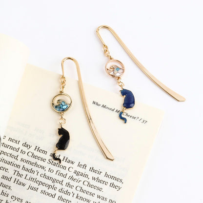 Close-up of gold cat bookmark with blue wave charm hanging outside book page, whimsical page marker detail