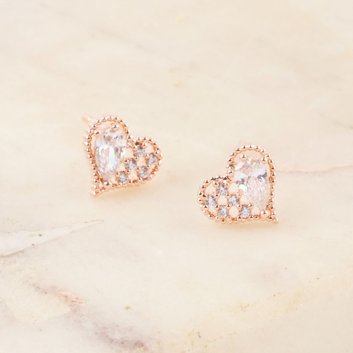 Close-up of rose gold heart-shaped stud earrings with cubic zirconia details, displayed on a soft marble surface.