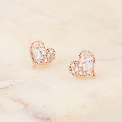 Close-up of rose gold heart-shaped stud earrings with cubic zirconia details, displayed on a soft marble surface.