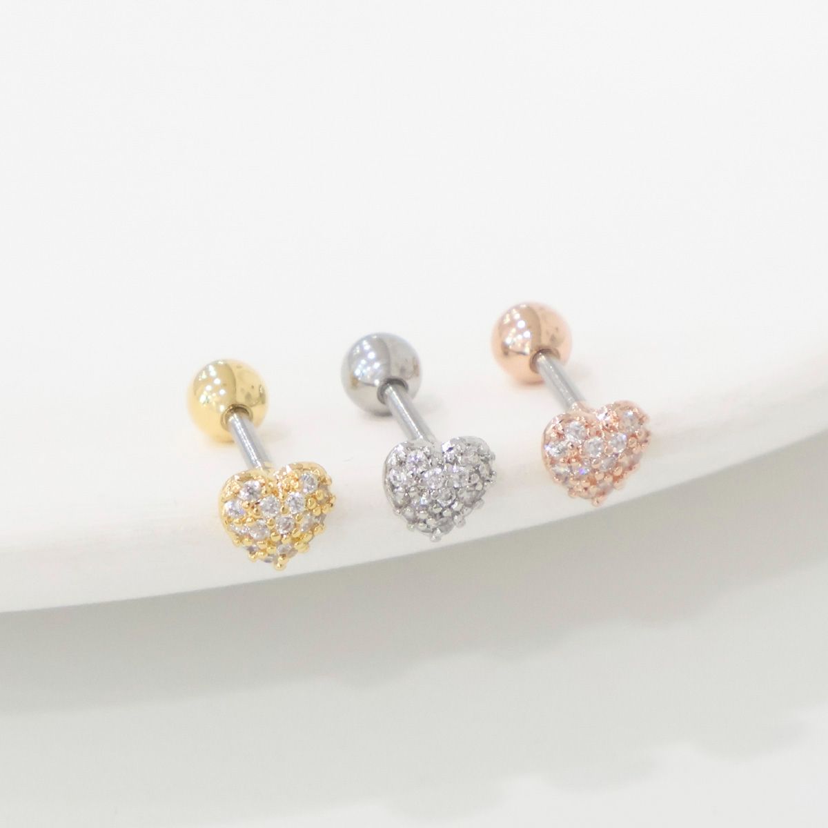 Three pavé heart barbell earrings in gold, silver, and rose gold displayed on a white ceramic tray.