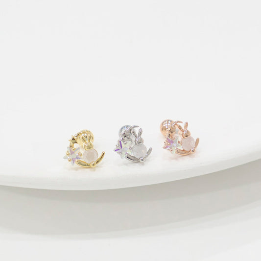 Barbell earrings in gold, silver, and rose gold, each shaped like a bunny sitting on a crescent moon with sparkling star details, arranged in a row on a white curved plate.