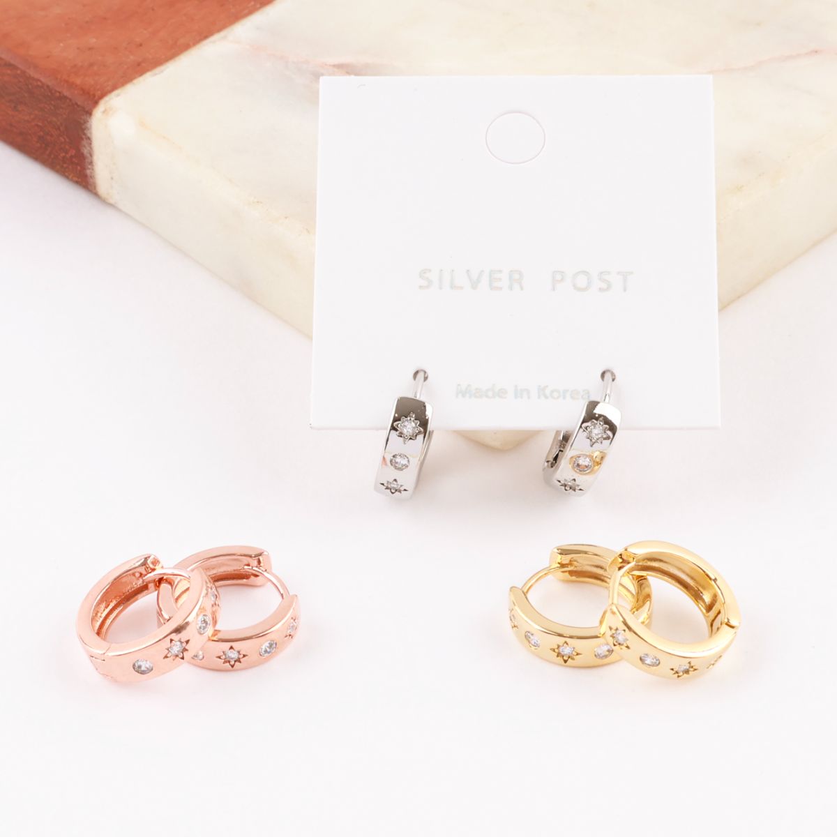 Set of three star-detailed huggie hoop earrings in silver, rose gold, and yellow gold displayed on card and marble block.