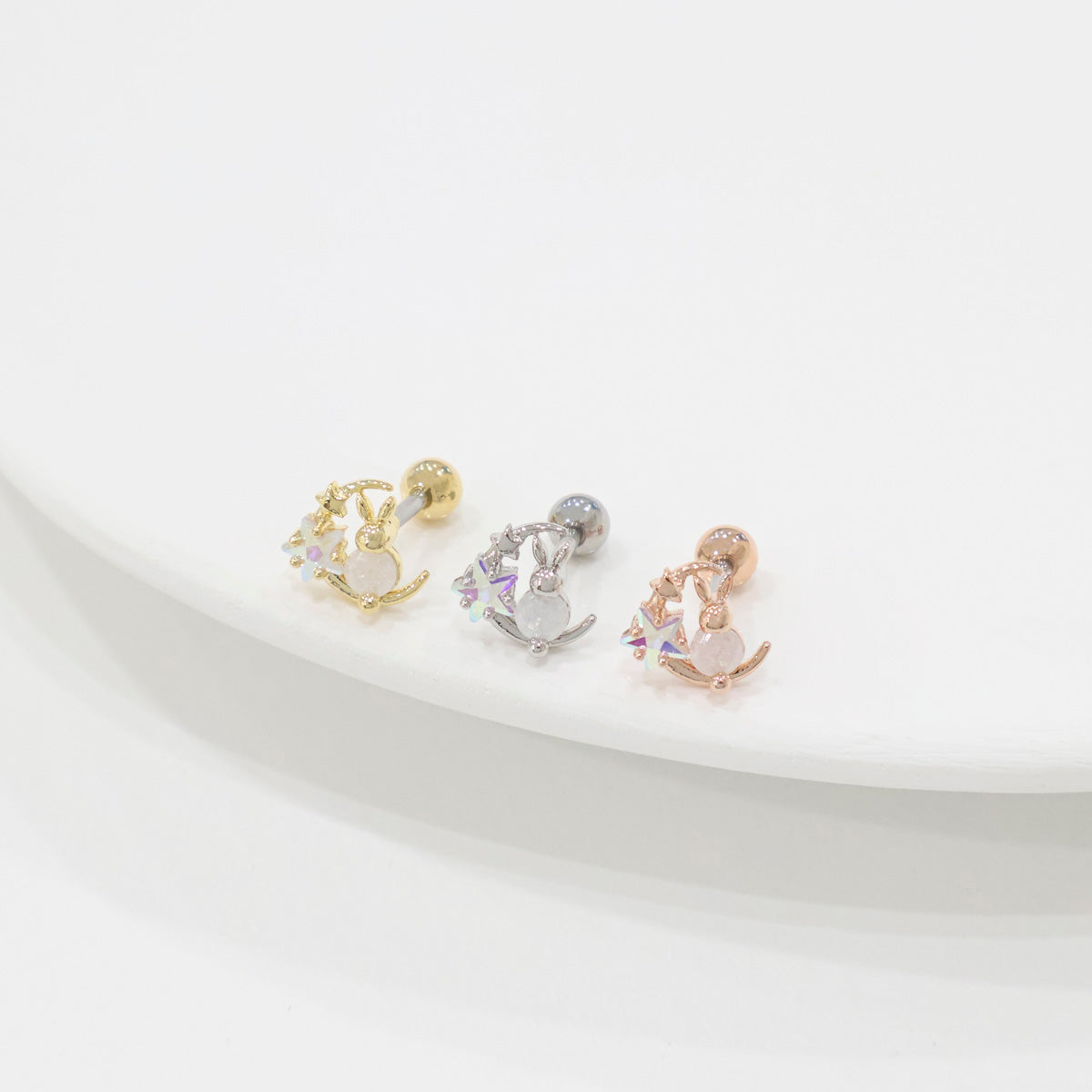 Three bunny moon barbell earrings viewed from a higher angle, showing gold, silver, and rose gold variants neatly lined up on a soft white surface.