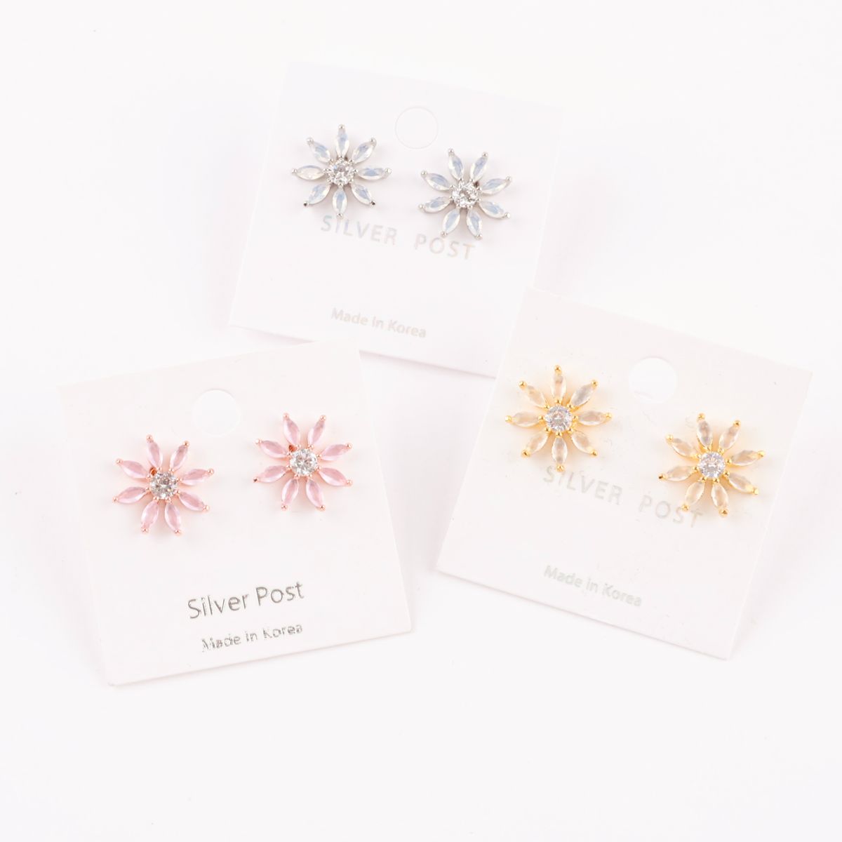 Set of three color variants of daisy-like earrings (gold, pink, and silver) arranged in front of their matching earring cards.