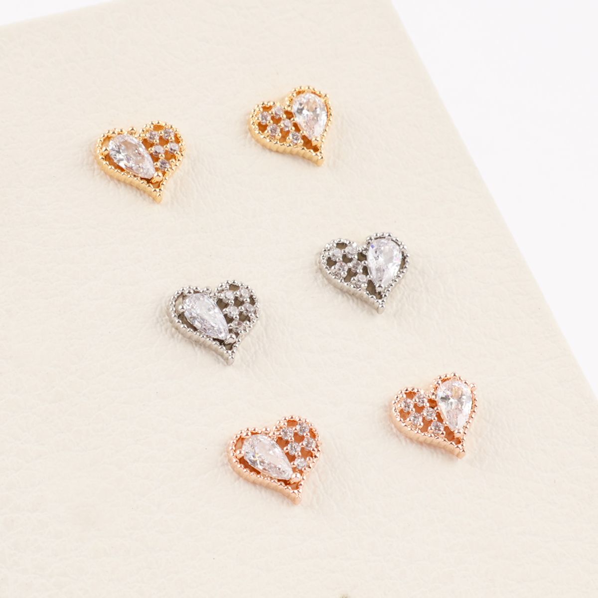 Three color variations of heart-shaped cubic stud earrings in gold, silver, and rose gold, arranged in pairs on a cream textured background.
