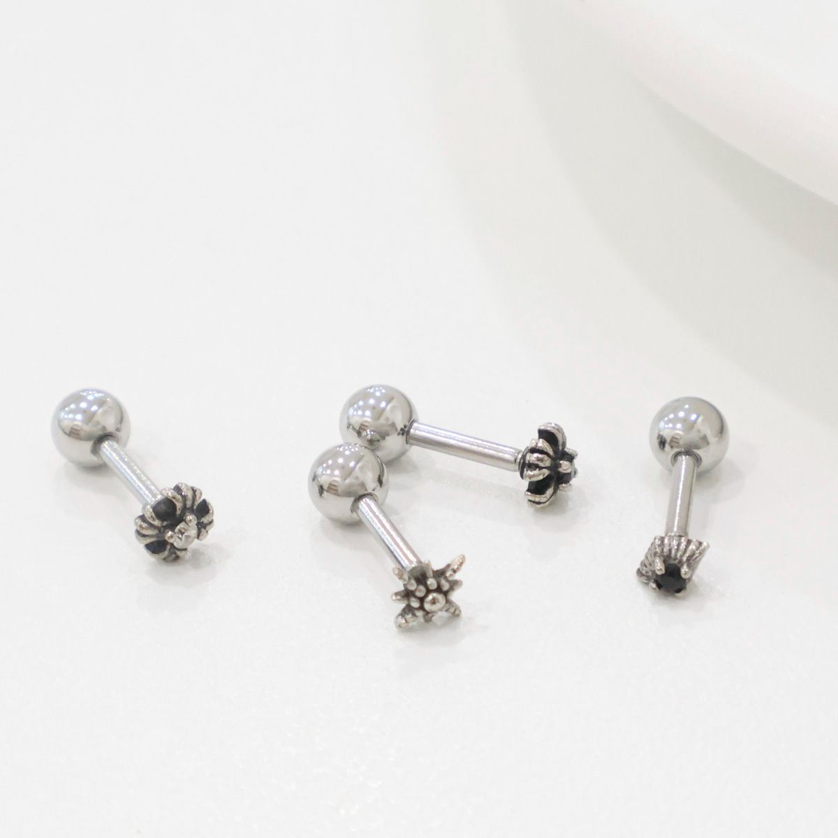 Angled flatlay of four stainless steel barbells on glossy white surface, showing the charms from a top view. Perfect for minimalist or edgy piercings.