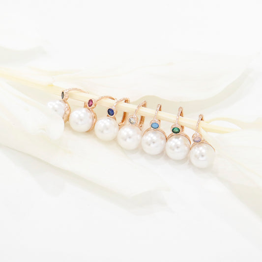 Close-up of 8 pearl earrings in rose gold hooks with colorful cubic stones, laid across white leaf props.