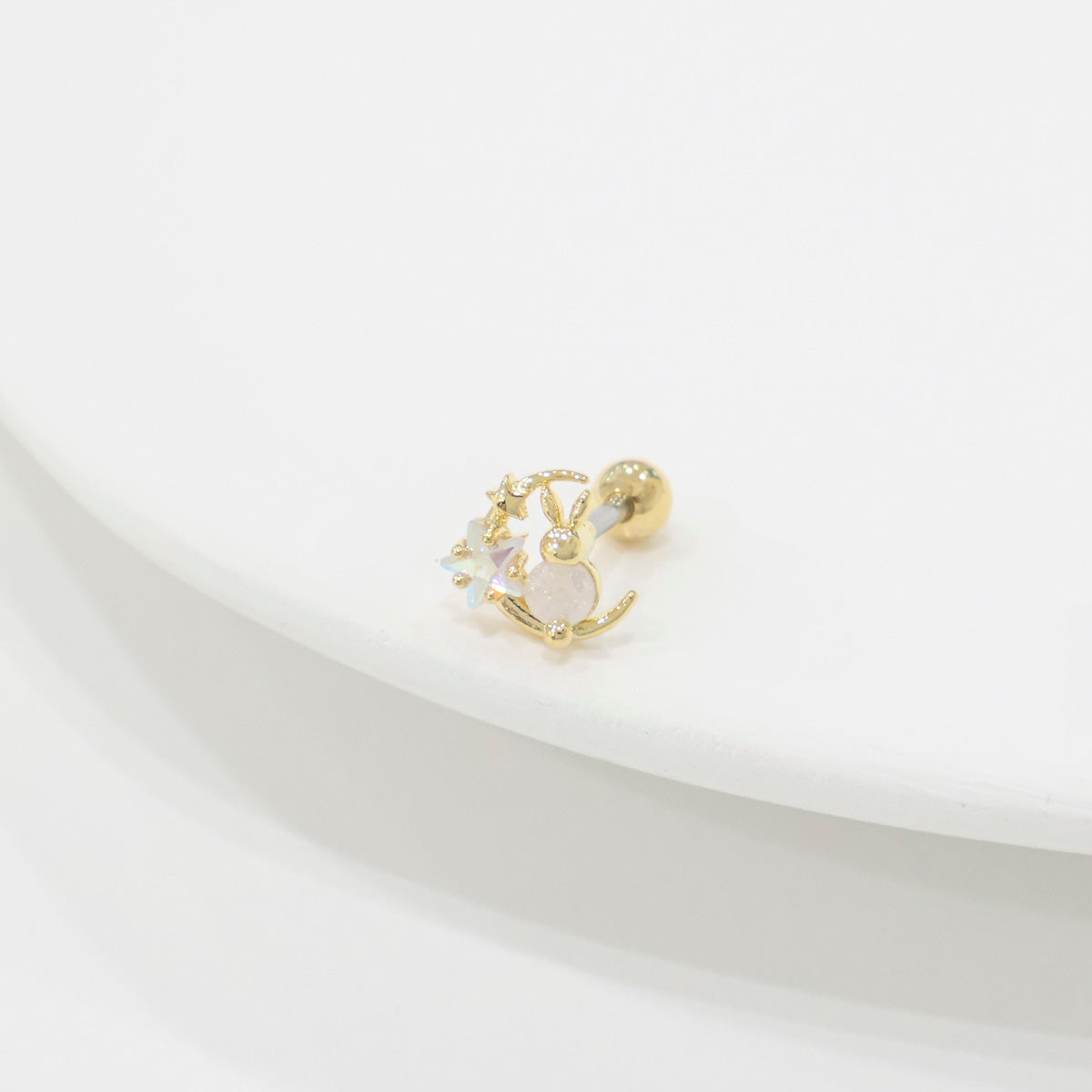 Single gold bunny moon barbell earring placed on a curved white plate, shot with soft shadows to highlight the iridescent stars and moon charm.