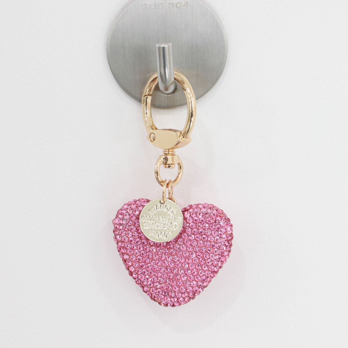 Close-up of a pink rhinestone heart keychain hanging on a silver wall hook.
Gold ring and round tag attached above the heart charm.