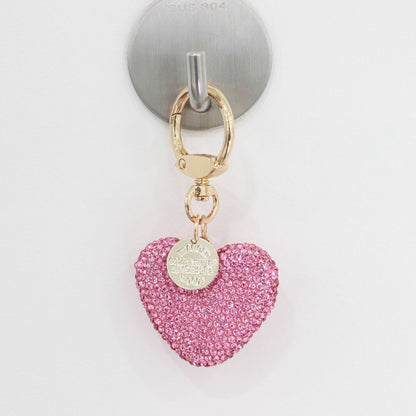 Close-up of a pink rhinestone heart keychain hanging on a silver wall hook.
Gold ring and round tag attached above the heart charm.