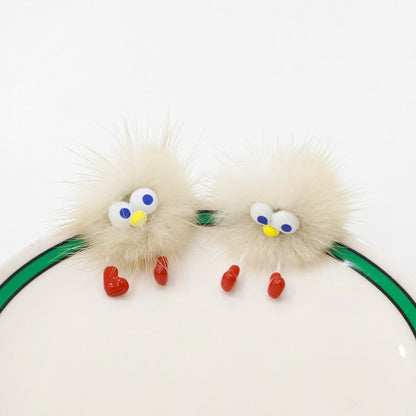 “Beige fluffy earring with googly eyes and red feet placed on a white plate with a green stripe