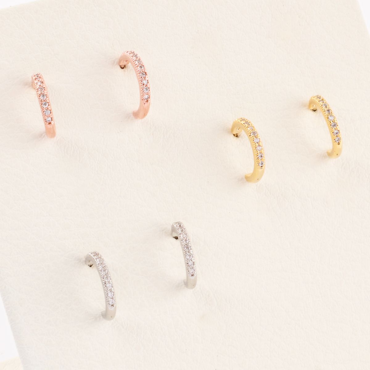 Six pairs of cubic zirconia half hoop earrings neatly arranged on a cream-colored velvet display, in silver, gold, and rose gold.