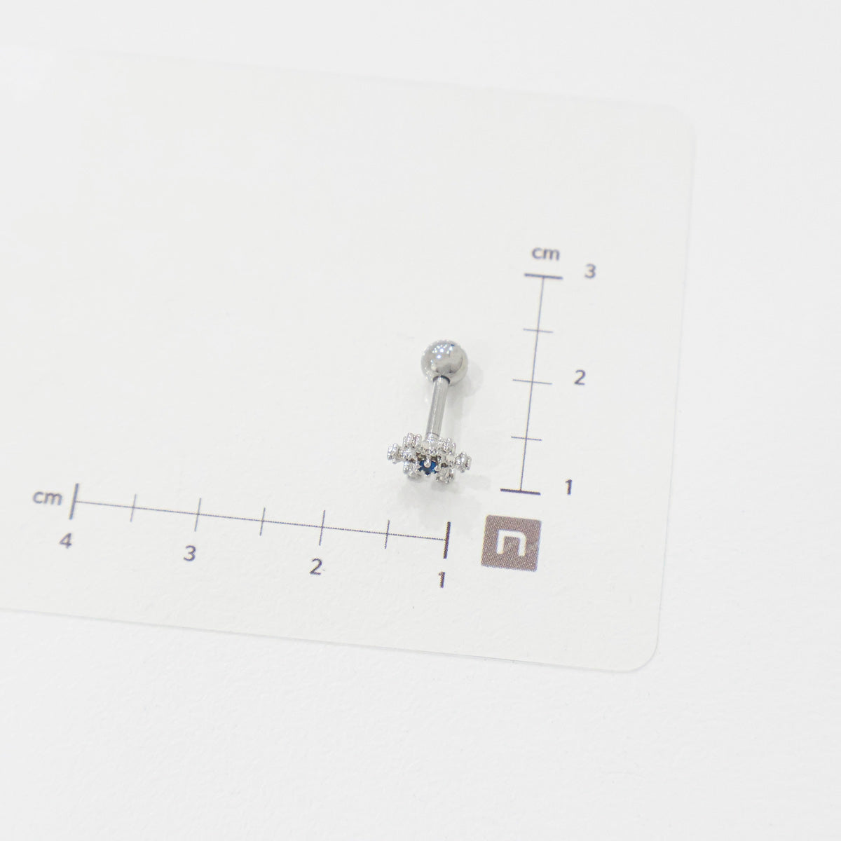 Snowflake barbell piercing with blue center stone placed on a measurement chart, showing approx. 1.3cm width.