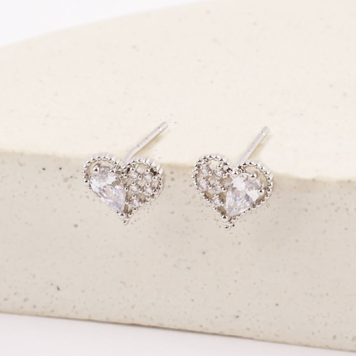 White gold heart stud earrings featuring pear-cut cubic stones, standing upright against a curved stone-textured backdrop.