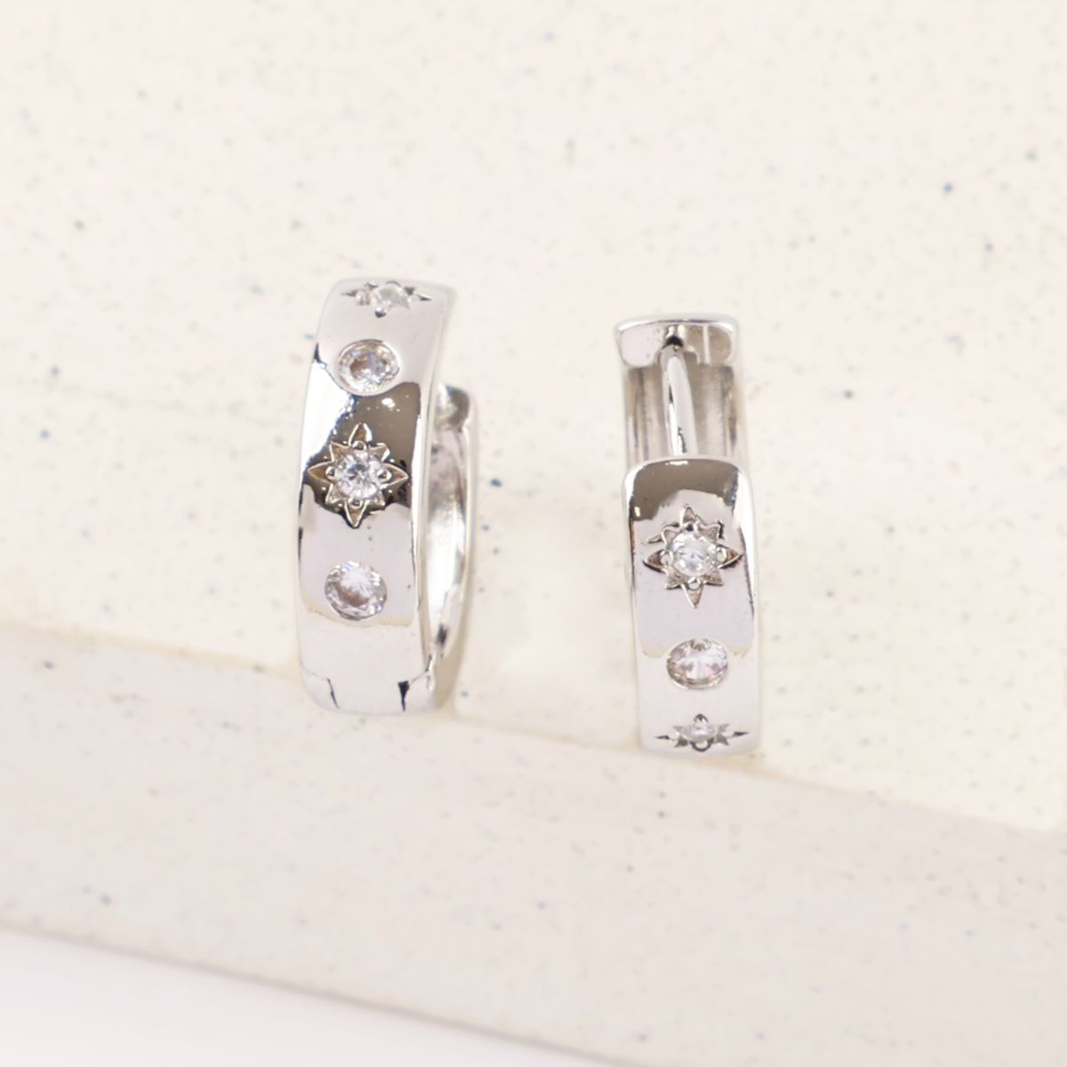 Close-up of silver huggie earrings with cubic zirconia and engraved stars on light speckled stone.