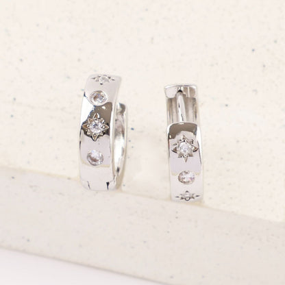 Close-up of silver huggie earrings with cubic zirconia and engraved stars on light speckled stone.