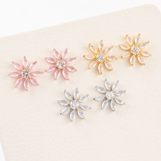 Six flower-shaped earrings in gold, pink, and silver tones displayed on a cream-colored textured surface.