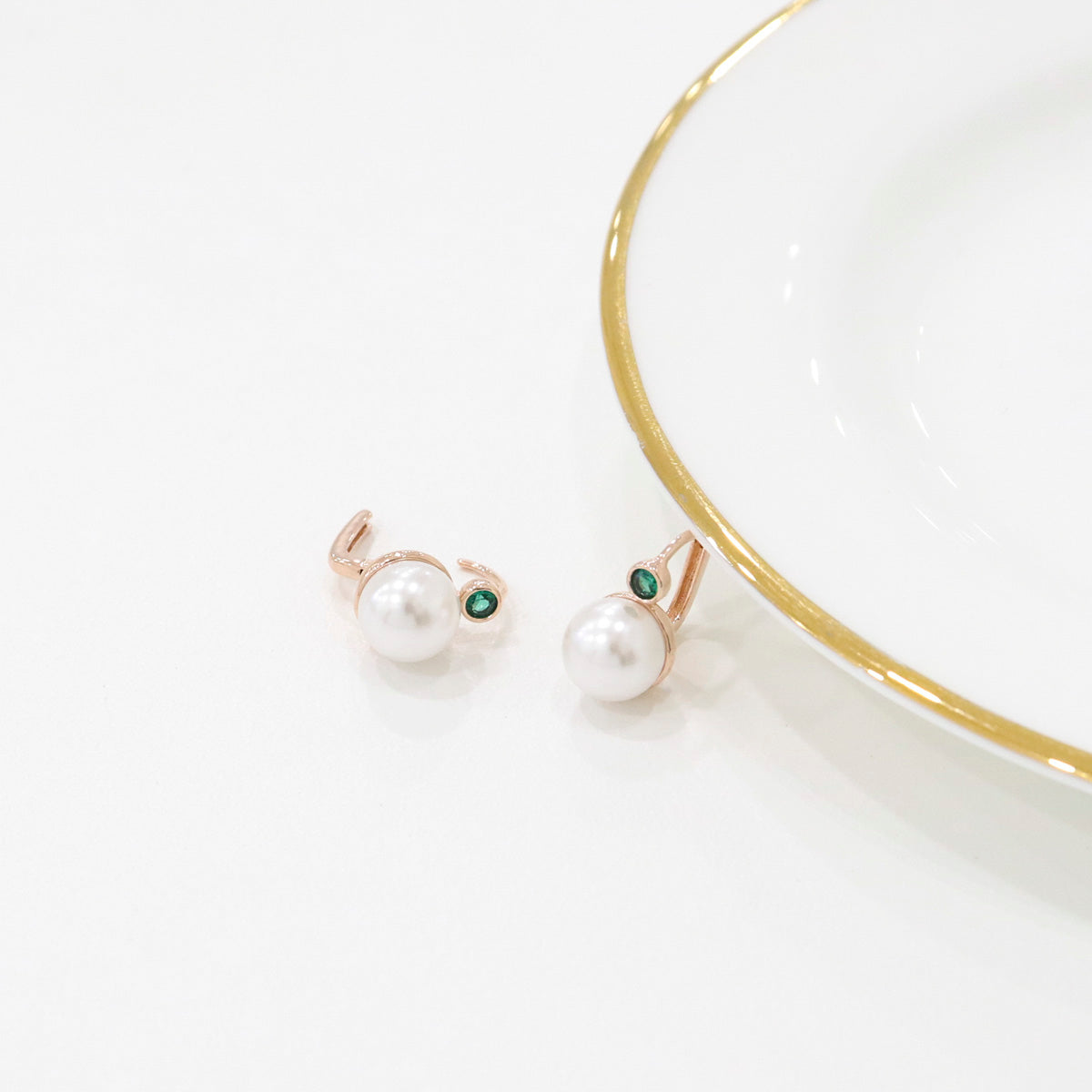 A pair of green-stone pearl drop earrings placed beside a white and gold-rimmed ceramic plate.