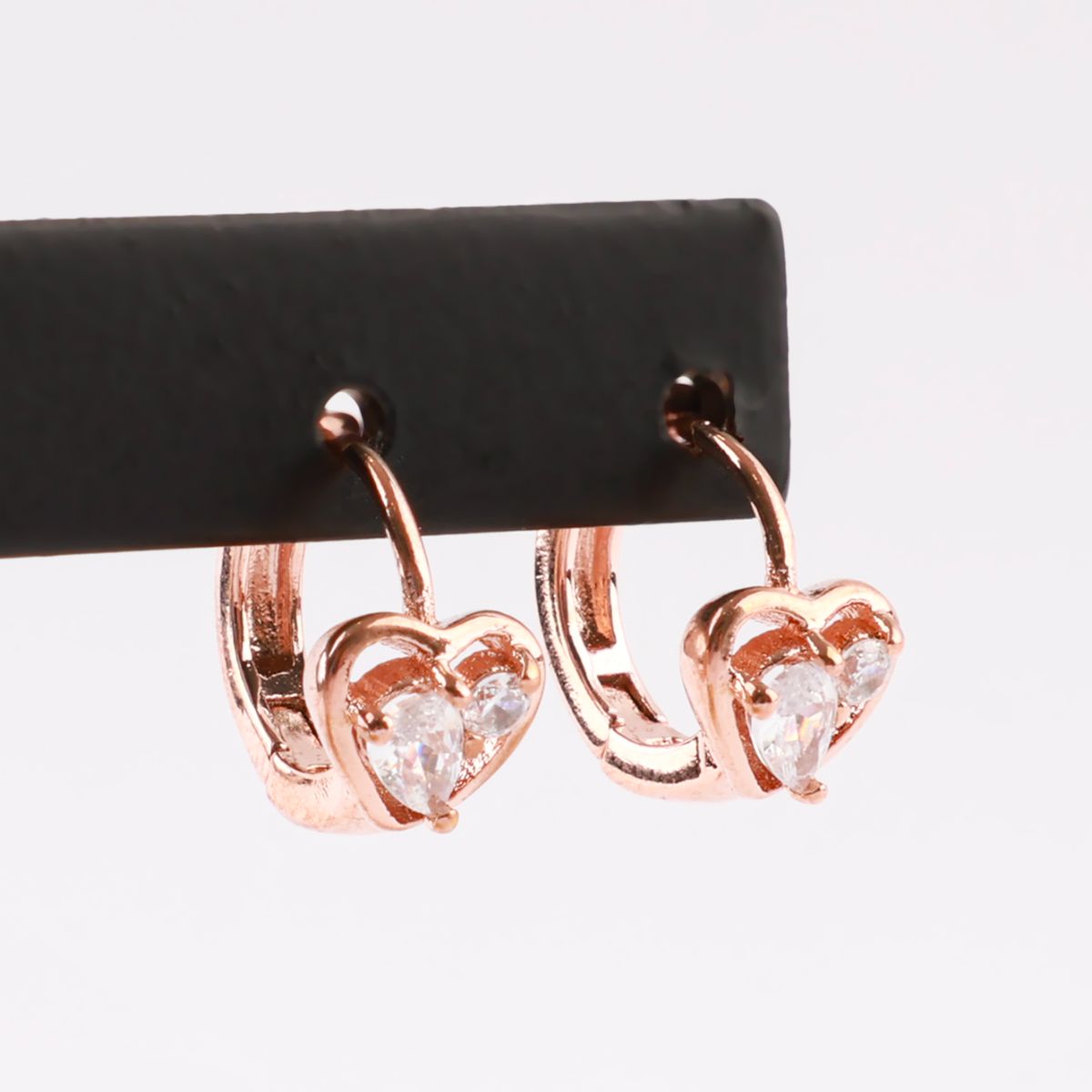 Rose gold heart cubic zirconia huggie earrings hanging from a black earring display bar, showing front view.