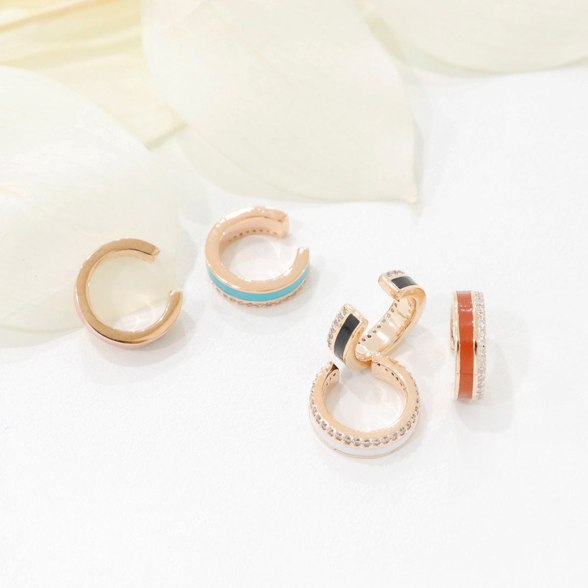Mixed enamel ear cuffs scattered on white surface with soft petal background — turquoise, black, brown