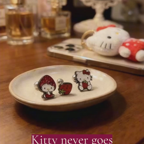 Hello Kitty strawberry surgical steel barbell earring set featuring three enamel designs on white background, strawberry costume Kitty, mini strawberry fruit, and sitting Kitty with strawberry, black metal borders with red enamel and green accents, 16G surgical steel barbells with ball studs, hypoallergenic piercing jewelry, Korean handmade Sanrio character piercings